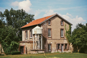 Vue de la maison adossée au mur de clôture nord, à ouvertures encadrées de briques, accostée d'un puits surmonté d'une réserve d'eau métallique. © Région Bourgogne-Franche-Comté, Inventaire du patrimoine
