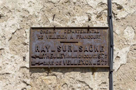 Plaque signalétique en fonte apposée sur la maison à l'angle de la Grande Rue et de la rue Sainte-Anne : CHEMIN DEPARTEMENTAL / DE VELLEXON A FRANCOURT / RAY-SUR-SAONE / THEULEY LES LAVONCOURT 5k7 / GARE DE VELLEXON 2K7. © Région Bourgogne-Franche-Comté, Inventaire du patrimoine