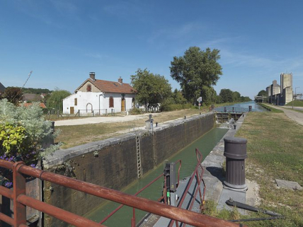 Panorama du site de l'écluse 113 du versant Yonne, dite de Cheny à Migennes, pris d'aval, du pont sur écluse. De gauche à droite : site d'écluse, silos. Le bief est vide : on voit la maçonnerie des bajoyers. © Région Bourgogne-Franche-Comté, Inventaire du patrimoine