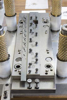Atelier de fabrication de l'outillage : une étampe progressive, avec une bande métallique témoin (à gauche). La bande de métal avance progressivement dans l'étampe et à chaque coup de presse, une opération est effectuée. © Région Bourgogne-Franche-Comté, Inventaire du patrimoine Atelier de fabrication de l'outillage : une étampe progressive, avec une bande métallique témoin (à gauche). La bande de métal avance progressivement dans l'étampe et à chaque coup de presse, une opération est effectuée. © Région Bourgogne-Franche-Comté, Inventaire du patrimoine