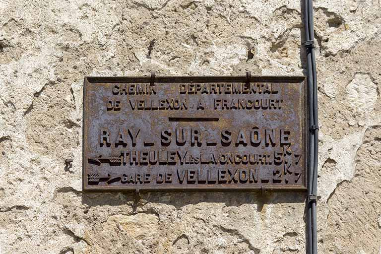 Plaque signalétique en fonte apposée sur la maison à l'angle de la Grande Rue et de la rue Sainte-Anne : CHEMIN DEPARTEMENTAL / DE VELLEXON A FRANCOURT / RAY-SUR-SAONE / THEULEY LES LAVONCOURT 5k7 / GARE DE VELLEXON 2K7. © Sonia Dourlot / Région Bourgogne-Franche-Comté, Inventaire du patrimoine - 2015