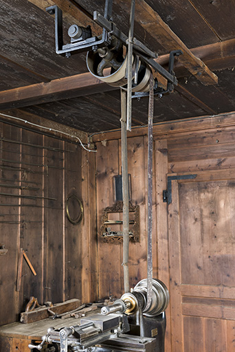 Exemple de disposition : machine à fraiser (rangée de droite). Le levier vertical au plafond permet de changer de poulie la courroie motrice (absente) tandis qu'une deuxième courroie transmet l'énergie à la machine, équipée d'une poulie étagée. © Sonia Dourlot / Région Bourgogne-Franche-Comté, Inventaire du patrimoine - 2017