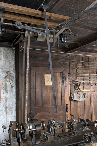 Exemple de disposition : machine à décolleter (rangée de droite). Le levier vertical au plafond permet de changer de poulie la courroie motrice (à droite) tandis qu'une deuxième courroie transmet l'énergie à la machine, équipée d'une poulie étagée. © Sonia Dourlot / Région Bourgogne-Franche-Comté, Inventaire du patrimoine - 2017